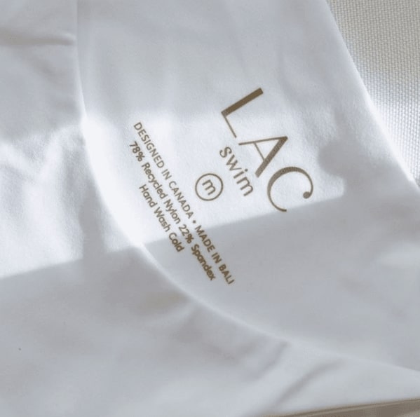 Close up of a LAC swimsuit bottom in white. The logo says LAC Swim and explains that it is designed in Canada and made in Bali.