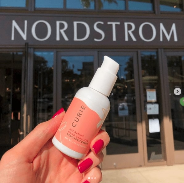 Close up of a pink bottle of curie hand sanitizer. In the background are the front doors to a store with a big sign above that says _Nordstrom_