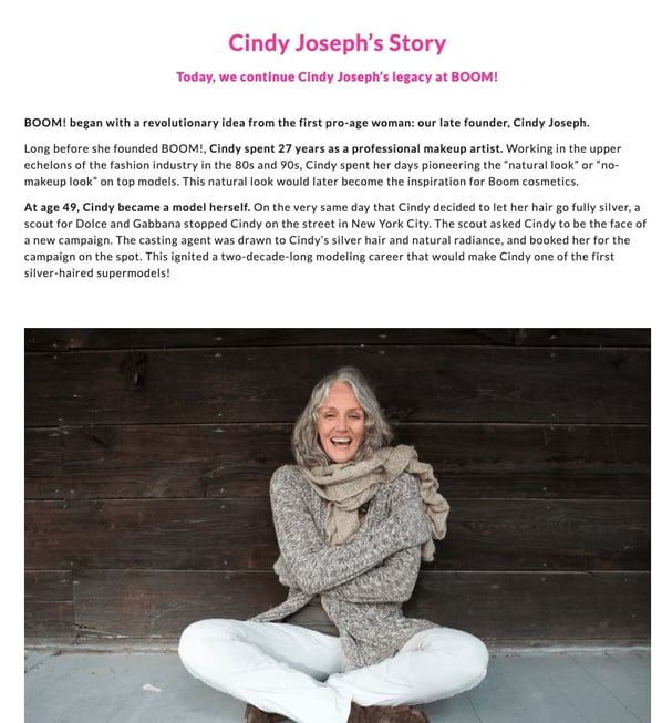 Screenshot of BOOM! By Cindy Josephs about page. There is a picture of Cindy sitting on the ground. She is wearing white pants and a grey sweater. She is hugging herself and smiling
