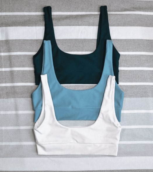 Three LAC Swim tops stacked on top of each other. One is dark green_ one is light blue_ and one is white. They are on top of a striped grey background