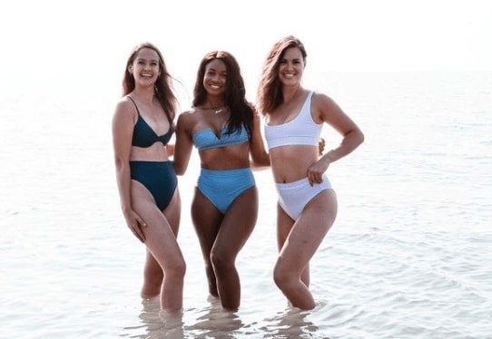 Three women standing in a lake wearing LAC swim suits. The left suit is dark green_ the middle is light blue and the right is white. They have their arms around each others backs_ smiling to the camera