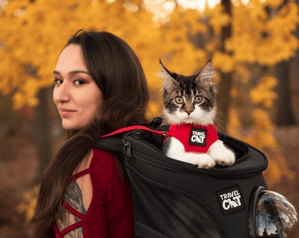 Woman with long brown hair in a red shirt wearing a black backpack. There is a white and grey cat popping its head out of the top of the backpack