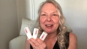 Woman with white hair holding three lipsticks by Boom! By Cindy Joseph. The background is a white wall.