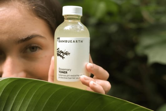 close up of a bottle of Bambu Earths rosemary toner. Behind the bottle is a woman looking at it. Theres a large leaf covering half her face and part of the bottle